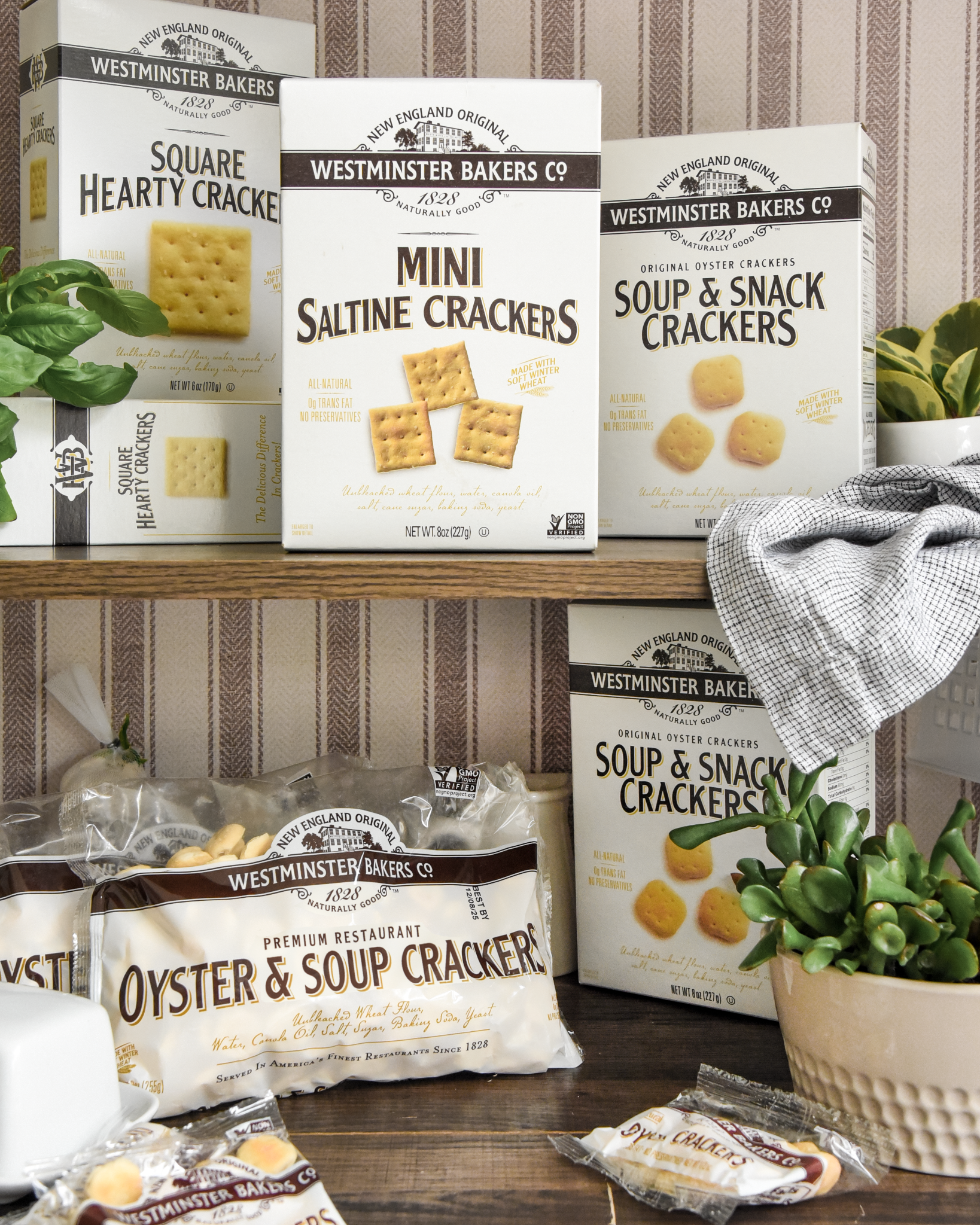 Assorted boxes and bags of Westminster Bakers Co. crackers on a wooden surface with plants and a checkered cloth.