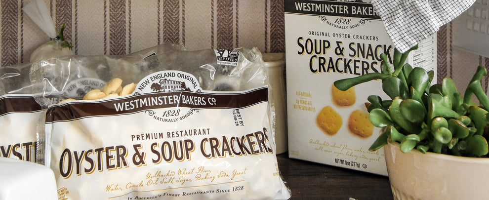 Packages of Westminster Bakers Oyster & Soup Crackers on a wooden surface with a plant in the background.