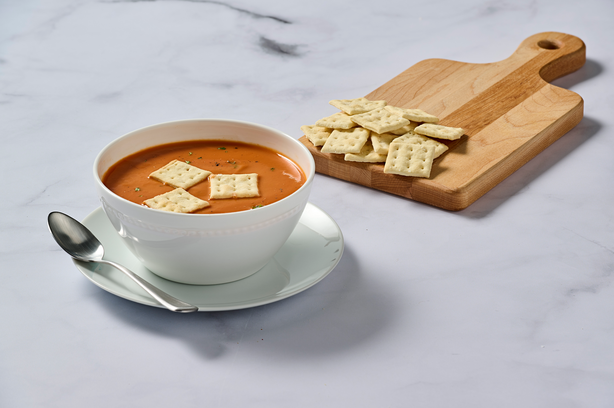 Tomato soup in a white bowl with crackers on a wooden board on a marble surface