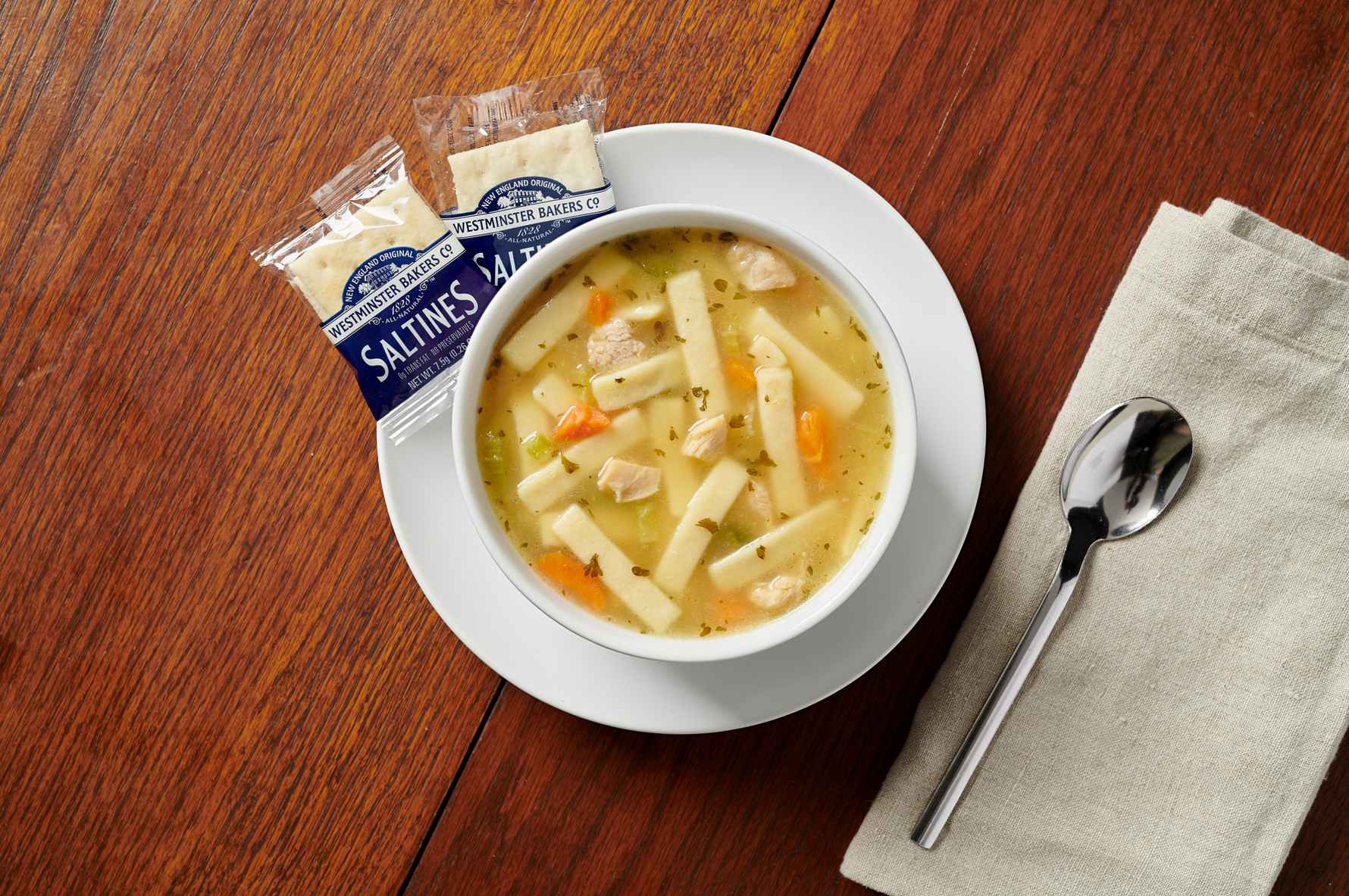 Bowl of soup with noodles and vegetables on a wooden table with a saltine cracker package.