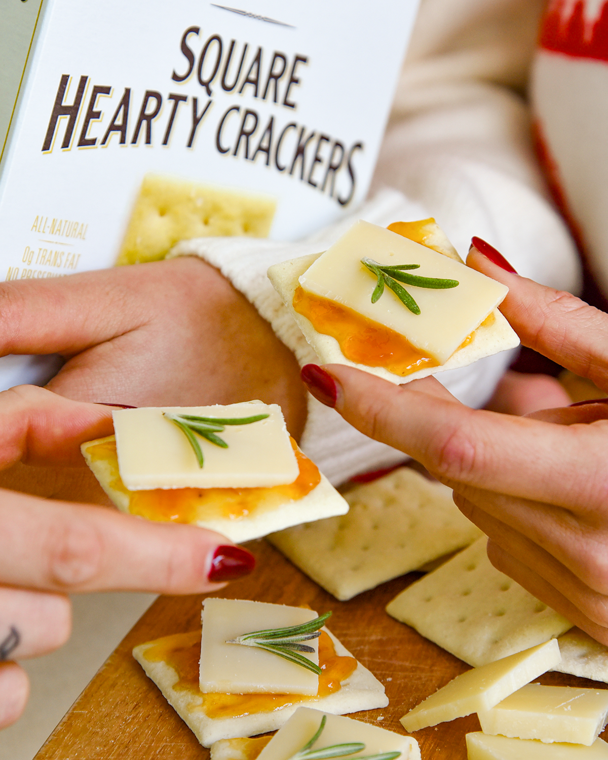 Square Hearty Crackers with cheese and rosemary being held by hands, with a box of crackers in the background.