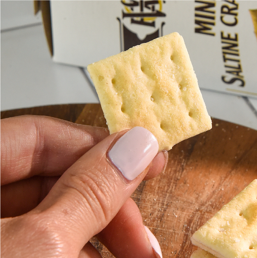 Hand holding a Saltine cracker with a box of crackers in the background