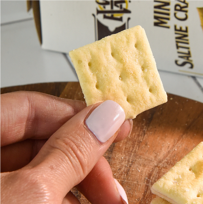 Hand holding a Saltine cracker with a box of crackers in the background